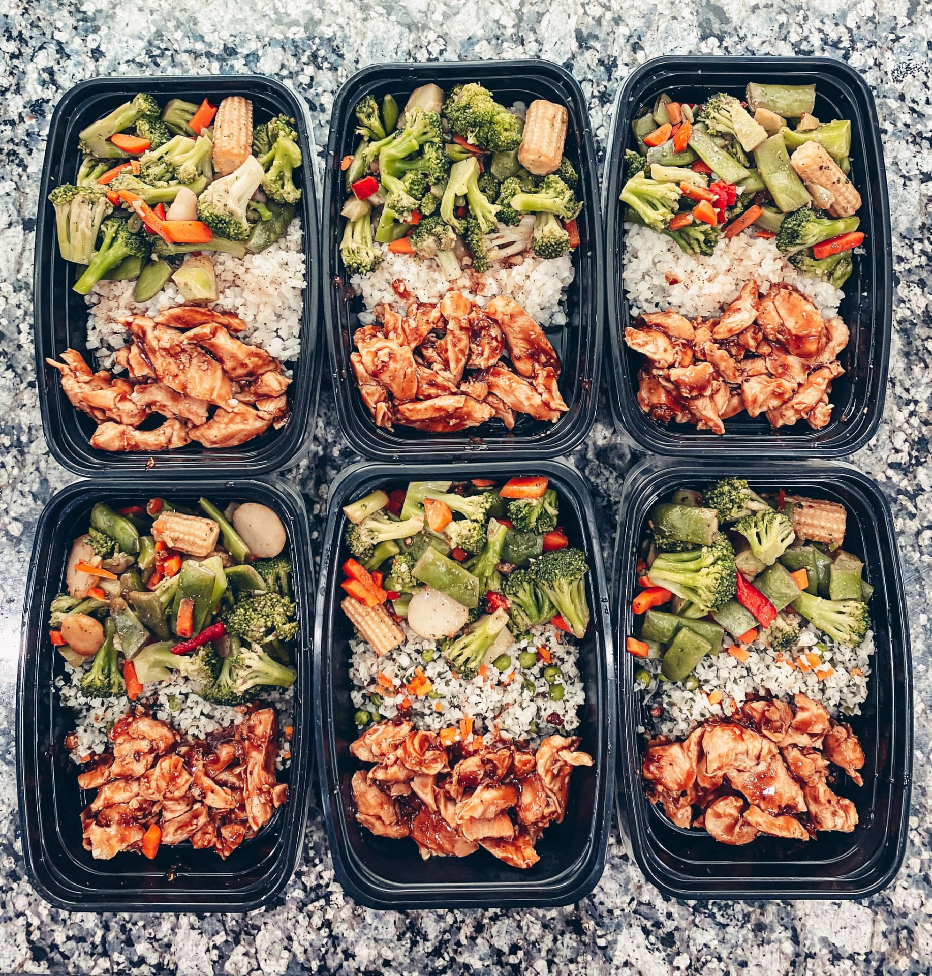 Six meal prep containers filled with rice, vegetables, and teriyaki chicken on a granite countertop